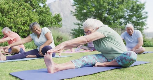 Diverse group of happy male and female seniors stretching on yoga mats in sunny garden, slow motion