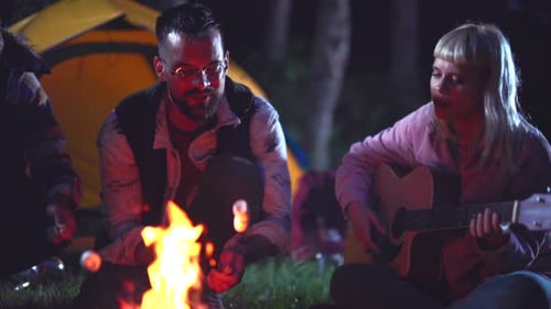 Happy multiracial friends having fun while camping together at night in the woods, roasting marshmal