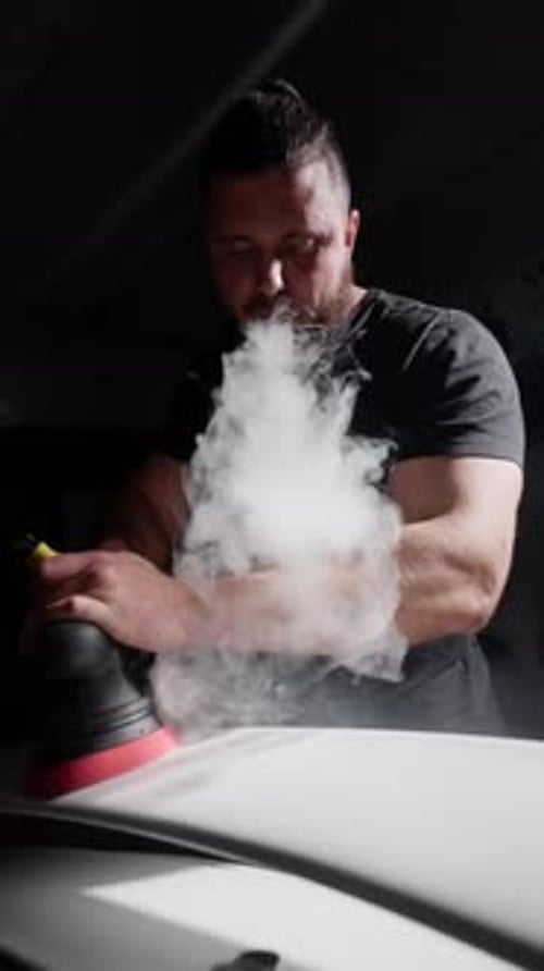 A Man Blows Smoke From an Ecigarette While Polishing a White Car