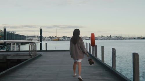 A figure strolls alone on a dock, with boats nearby and a gentle evening light reflecting off the wa