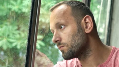 Melancholic man lost in thought gazing out window at rainy park in super slow motion