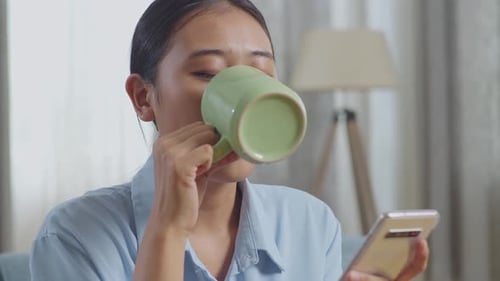 Close Up Of Asian Woman Drinking Coffee While Using Smartphone On Sofa In The Living Room