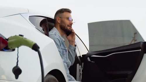 A Man Talking on the Phone While Charging Luxury Electric Car at Charging Station
