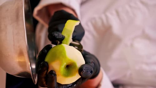 Vertical Video Closeup of a Chef's Hands in the Kitchen in a White Jacket Peeling Green Apple Into a
