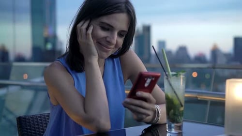 Young woman enjoys cocktail and texts on smartphone at luxury rooftop bar