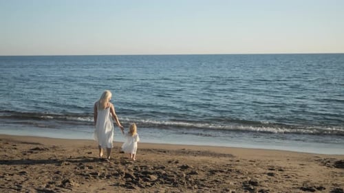 Mom and Little Daughter Holding Hands Walk Along the Beach to the Sea