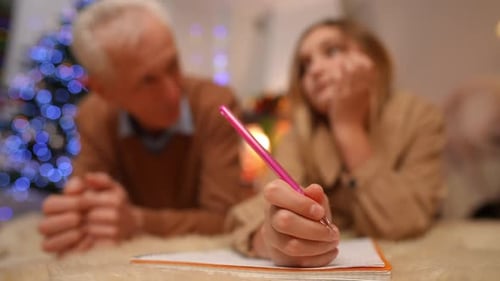 Closeup Teen Female Hand Writing Letter to Santa with Blurred Senior Man and Girl Talking at
