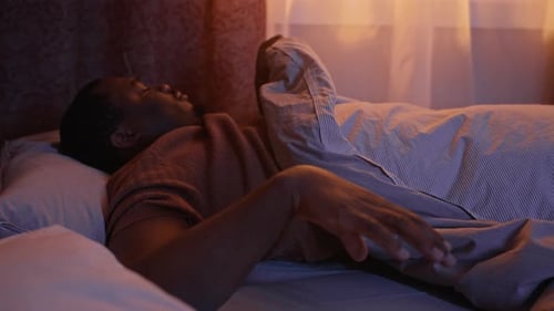 Schwarzer Mann hat Schlaflosigkeit und schaut nachts im Bett auf sein Smartphone
