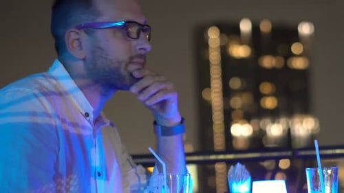 Handsome man impatiently checking phone and sipping cocktail waiting at rooftop bar night