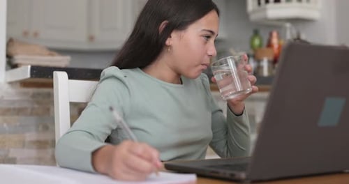Female kid drinking water while doing homework using laptop - Back to school