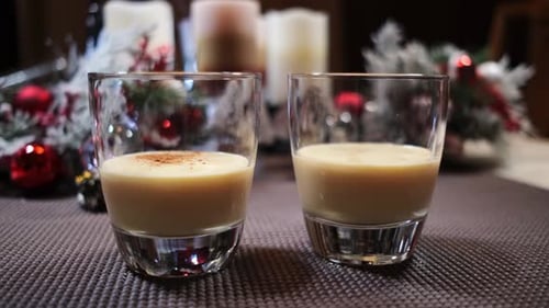 Two glasses of Christmas eggnog with ground cinnamon being added garnished with Christmas decoration