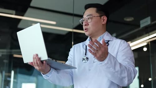 Male Physician Holding Laptop and Talking with Patient on Video Call in Office