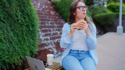 Frau beim Mittagessen mit einem Sandwich und Kaffee im Park mit einem Laptop