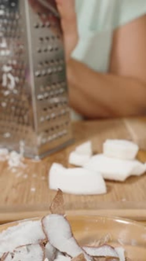 Vertical video. The Woman Grates Coconut Pieces, Turning Them into Shreds, Using a Metal Grater.