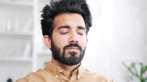Close up. Young handsome bearded man meditating with eyes closed while sitting on sofa in living