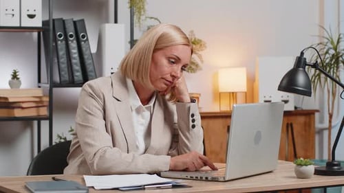 Bored Sleepy Mature Businesswoman Working on Laptop Yawns Leaning on Hand at Home Office Desk
