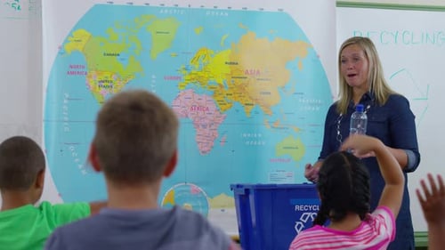 Teacher Giving Recycle Lesson In School Classroom And Student Goes Up Front To Help