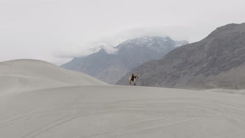 Um cavaleiro solitário atravessa as vastas dunas de areia do Paquistão com majestosas montanhas cobertas de neve