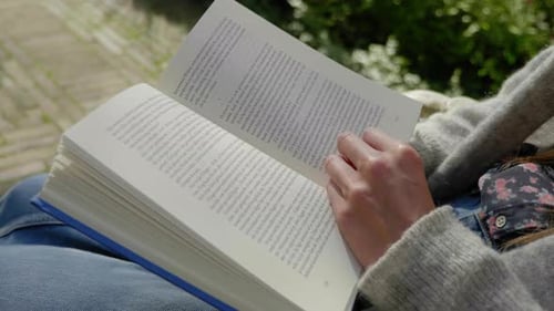 Slow motion close up shot of a book being read and by a young woman sitting, turning a page