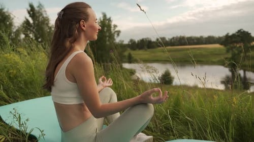 Woman Exercising Yoga Outdoors in Front of a Pond with Cloud Reflections in the Water