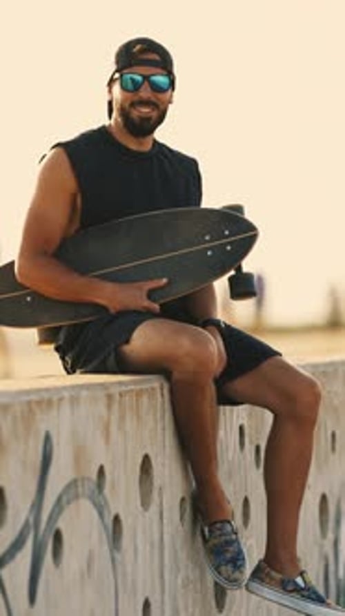 Man Sitting with Skateboard Wearing Sunglasses and Baseball Cap