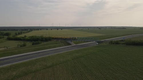 Ein gelber Personenzug fährt über eine Stahlbrücke über eine Autobahn in einer weiten ländlichen Landschaft.
Das