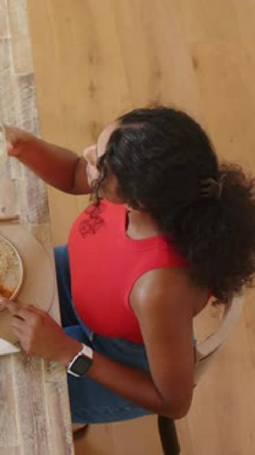 Vertical video: Sipping from glass with straw, woman in red top eating meal with fork in kitchen