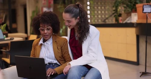 Two diverse female coworkers sitting together in a modern office collaborating on a business project
