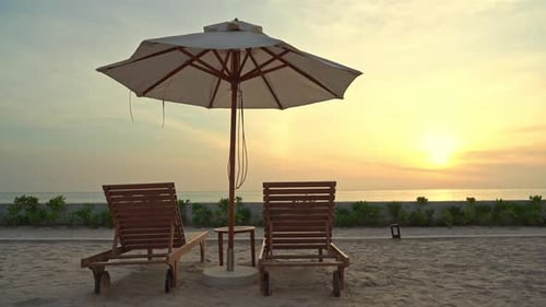 Close-up of two empty beach chairs and a shade umbrella on a sandy beach facing a colorful ocean sun