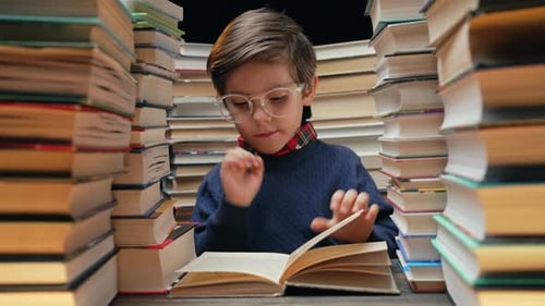 Cute Little Schoolboy in Glasses Reading Interesting Book in Library Between Stacks of Books