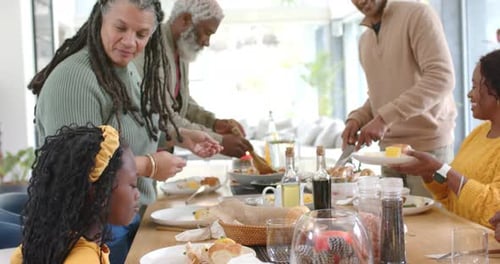 Diverse family passing dishes at wooden table when mature woman breaking bread for shared breakfast