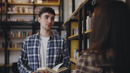 Young Man in Plaid Shirt Engaging in Conversation While Holding a Book in a Cozy Library Setting