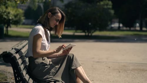 Young Woman Using Tablet Computer Sitting on Bench in City Park 30s