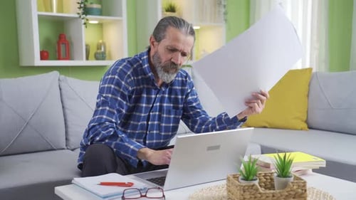 Man Reviewing Documents While Using Laptop at Home