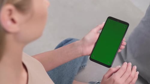 Eine Frau macht auf einem grünen Telefonbildschirm eine Wischbewegung nach links, während sie zu Hause auf der Couch sitzt. Swine