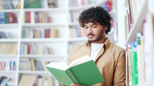 Student studying while reading a book while standing in campus library space. A handsome male
