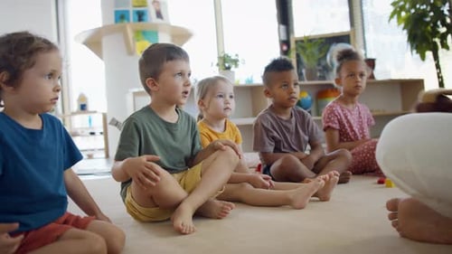Group Of Small Nursery School Children With Man Teacher Sitting On Floor Indoors In Classroom, Ha...
