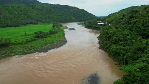 Drone view captures a winding brown river cutting between farmland and forested slopes, with