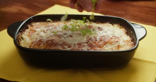 Chef Sprinkling Fresh Green Onions Over Baked Potatoes with Melted Cheese in a Cast Iron Baking Dish