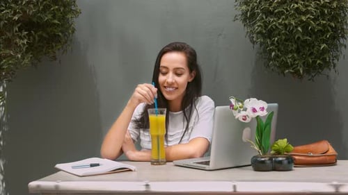 Smiling Woman Drinks Juice at Table with Laptop