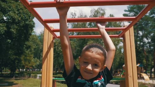 Young Boy Hang on Monkey Bars By His Hands to Exercise at Outdoor Playground Against the Sky in Slow
