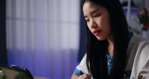 A young Asian woman taking extra lessons on a tablet while sitting at a table in living room.