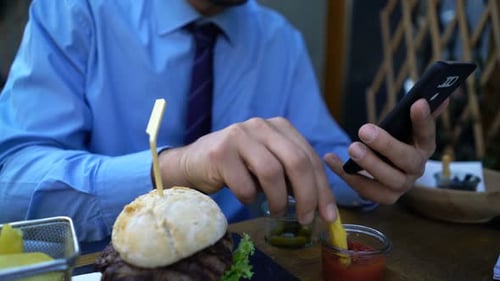 Businessman enjoys lunch break with burger and smartphone in the city cafe