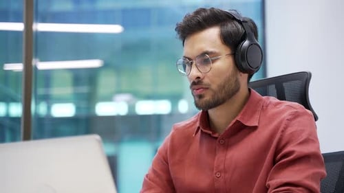 Focused IT specialist in wireless headphones works on a laptop sitting at workplace in business
