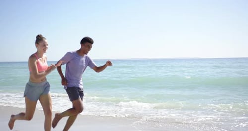 A young Caucasian woman and a biracial man, dressed in athletic wear, are jogging along the beach