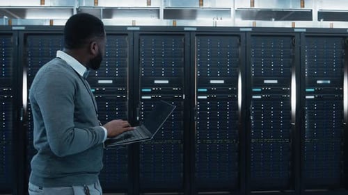 Black Data Center IT Technician Walking Through Server Rack Corridor with a Laptop Computer. He is