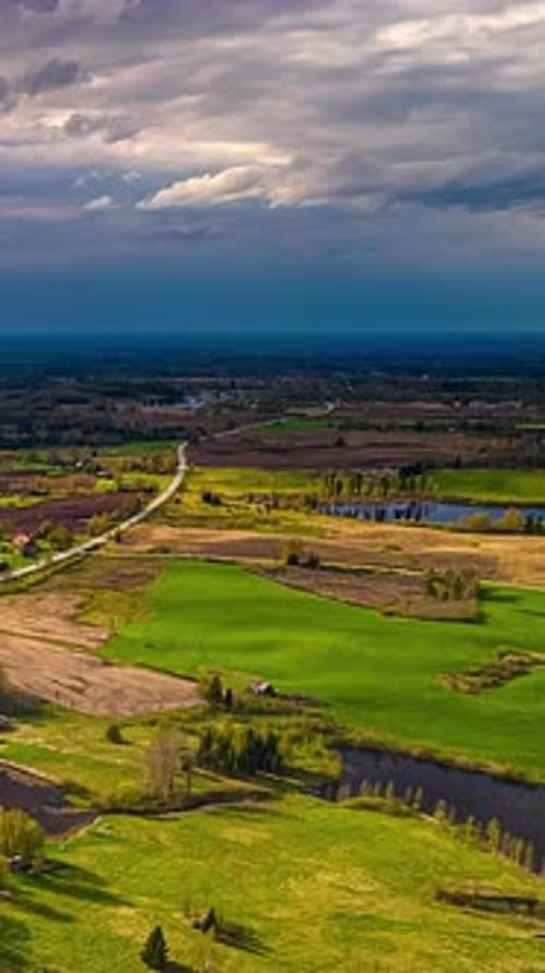 Vertical timelapse aerial view Latvia countryside, winding road, green meadows, river, dramatic sky