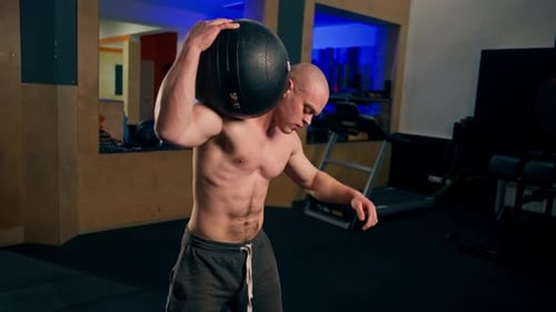 In the Gym Young Handsome Bald Trainer Lifts a Training Ball From the Floor Onto His Shoulders and