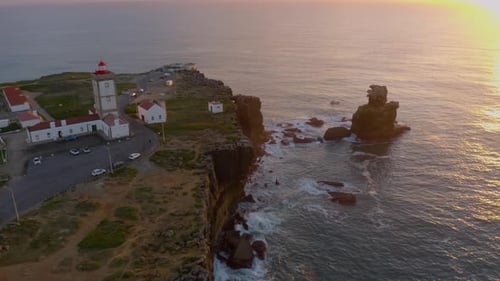 Aerial fly over coastal lighthouse with waves crashing on the cliffside.