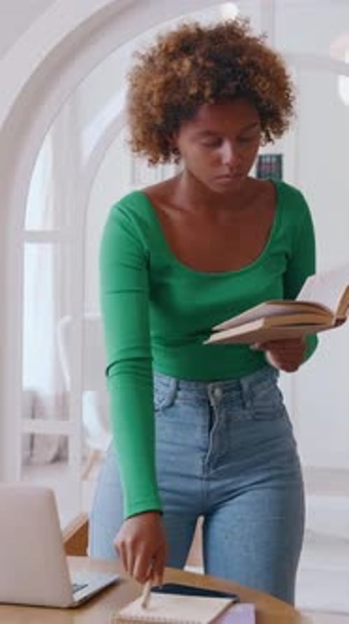 Young Woman Reading and Studying at a Home Office Standing By a Desk with Laptop